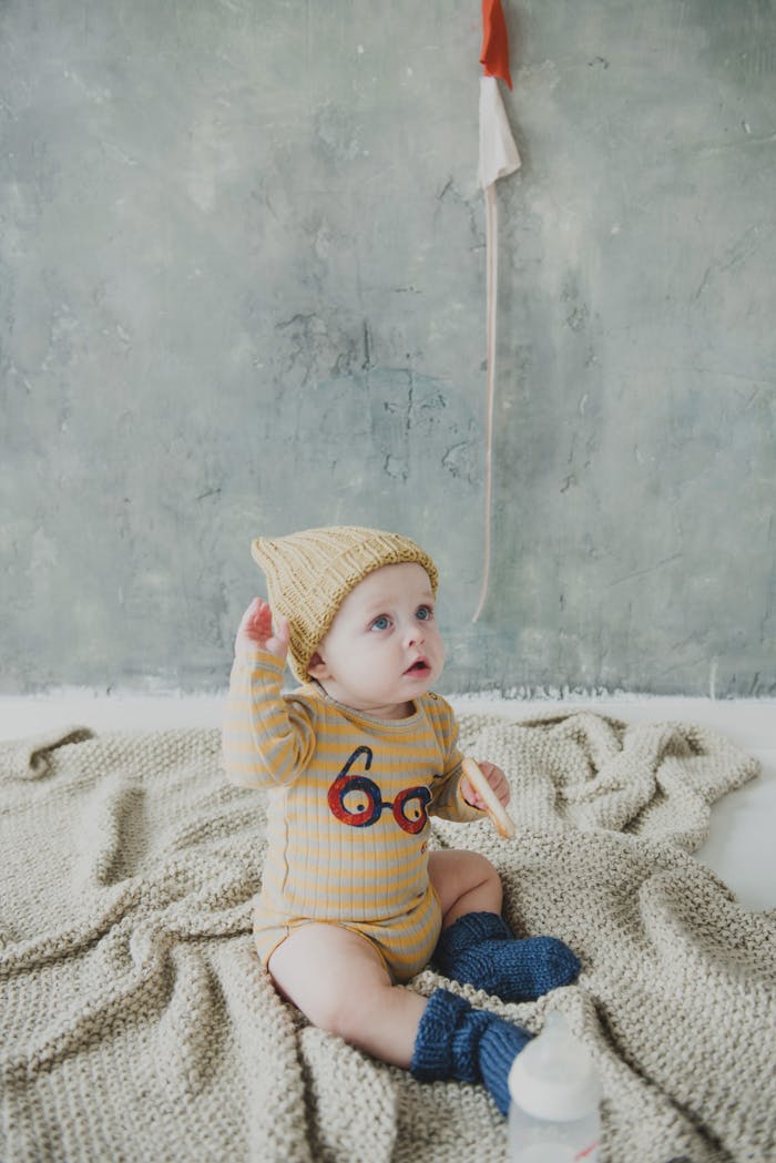 Charming baby in a knitted beanie and colorful onesie sitting indoors with a soft blanket.